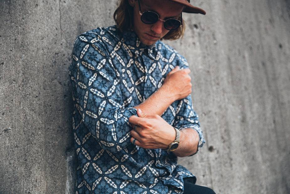 Man wearing ocean blue shirt with tiled kaleidoscope patterns and narrow collar adjusting sleeve against concrete wall