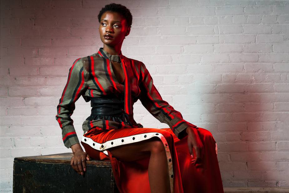 Woman wearing a black and red striped silk blouse with buckle collar and matching red button pants sitting on a wooden box against a white brick wall